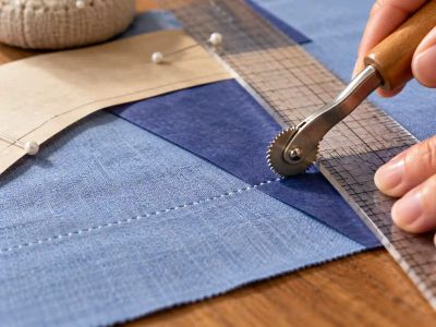 A serrated tracing wheel rolled along a clear ruler over blue dressmaker's carbon, leaving a dotted line on the fabric.