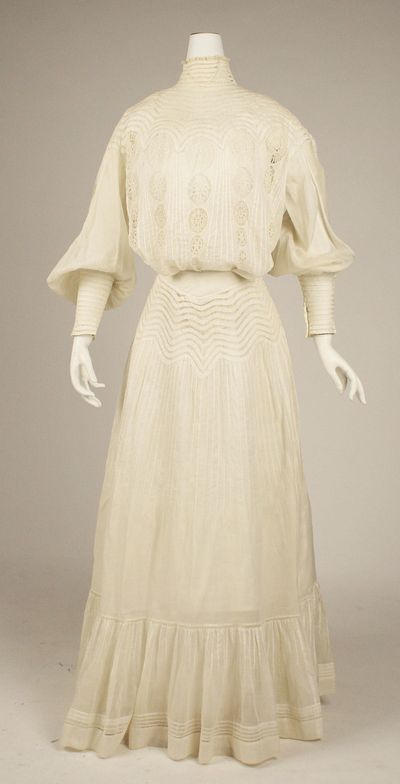 A white cotton day dress of 1903 on a dress form, with a high collar, a bodice pouched over the waist, rows of tucks and lace insertion, and a skirt that flares toward the hem.